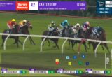 Group of galloping horses on a green turf racecourse with jockeys in colorful silks; on-screen graphics show race info and current placings/odds.