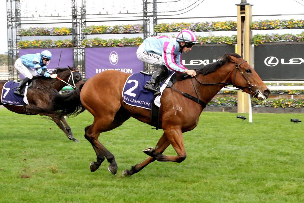 Two jockeys ride racehorses at Flemington, racing hard on a grassy track with banners and flowers in the background.