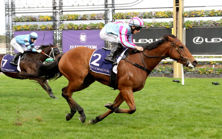 Two jockeys ride racehorses at Flemington, racing hard on a grassy track with banners and flowers in the background.