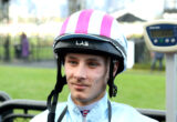 Young man wearing a pink-striped helmet with LAS logo, outdoors and smiling slightly.