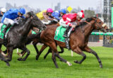 Group of thoroughbreds galloping at full speed during a horse race, jockeys in bright silks leaning forward on the track.