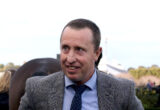 Man in a gray checkered blazer smiling outdoors at an event of people in the background.