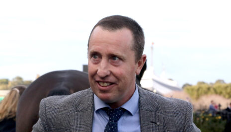 Man in a gray checkered blazer smiling outdoors at an event of people in the background.