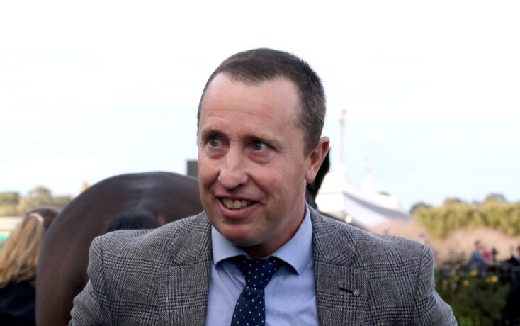 Man in a gray checkered blazer smiling outdoors at an event of people in the background.