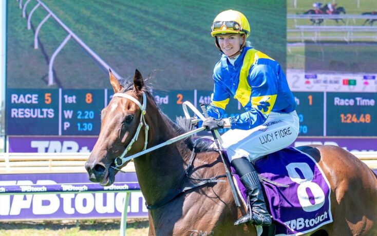 Jockey in blue and yellow silks rides a brown horse on a racecourse, scoreboard in the background.