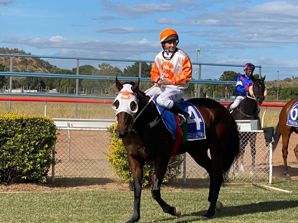 Jockey in orange and white silks riding a dark horse at a racetrack, with another rider in blue nearby and a blue sky above.