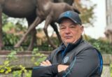 Man in a black jacket and cap taking a selfie in front of a large dinosaur statue in a park.