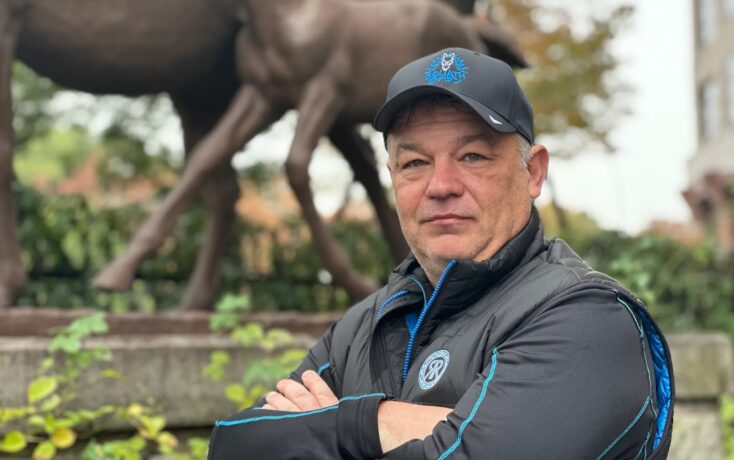 Man in a black jacket and cap taking a selfie in front of a large dinosaur statue in a park.