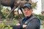 Man in a black jacket and cap taking a selfie in front of a large dinosaur statue in a park.