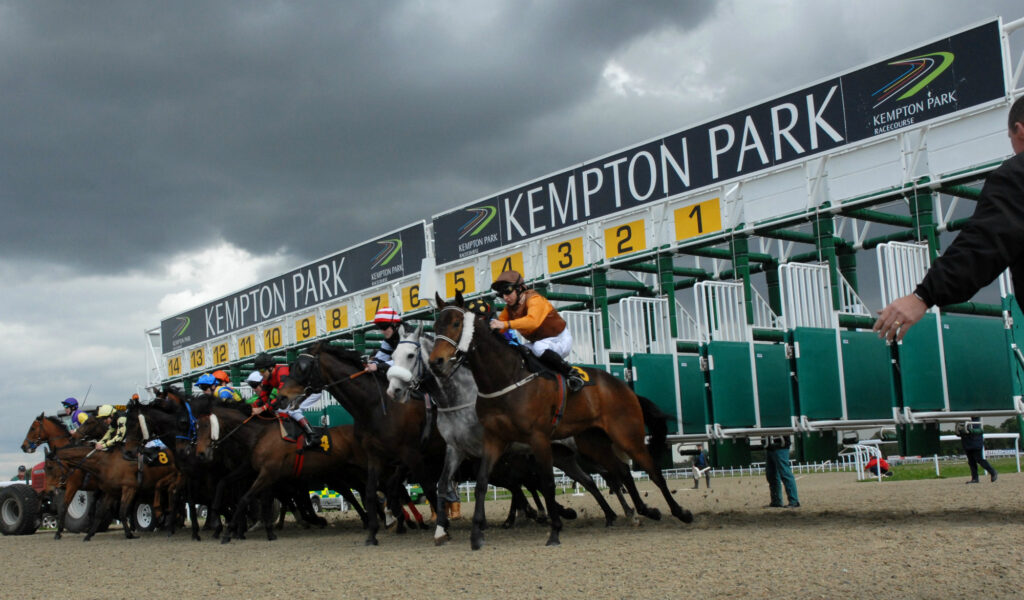 Horses and jockeys burst from the starting stalls at Kempton Park racecourse under dark clouds. ','Group of horse racers at the Kempton Park starting line, riders leaning forward as they speed off. ','Starting gates at Kempton Park with yellow numbered panels and jockeys on sprinting horses. ','Jockeys in colorful silks race from the gates at Kempton Park while a spectator stands nearby. ','Overhead view of a horse race launch at Kempton Park with a cloudy sky.