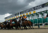 Horses and jockeys burst from the starting stalls at Kempton Park racecourse under dark clouds. ','Group of horse racers at the Kempton Park starting line, riders leaning forward as they speed off. ','Starting gates at Kempton Park with yellow numbered panels and jockeys on sprinting horses. ','Jockeys in colorful silks race from the gates at Kempton Park while a spectator stands nearby. ','Overhead view of a horse race launch at Kempton Park with a cloudy sky.