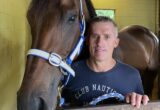 Man posing with a brown horse in a stable, horse wearing a blue and white halter, both facing the camera over a stall railing