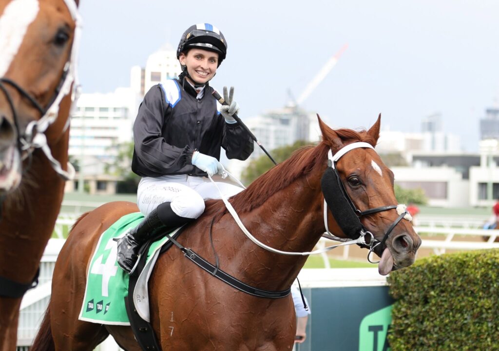 Leah Martyn | Just Horse Racing Smiling jockey in black-and-blue racing gear rides a chestnut horse on a city racecourse backdrop.