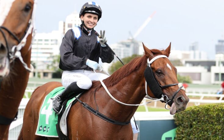 Smiling jockey in black-and-blue racing gear rides a chestnut horse on a city racecourse backdrop.