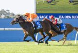Two jockeys race on a grassy turf, close together as their horses push toward the finish line, one in orange silks and the other in red and white.