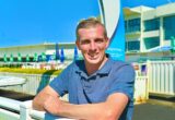 Smiling man in a blue polo leaning on a white railing at an outdoor aquatics facility on a sunny day