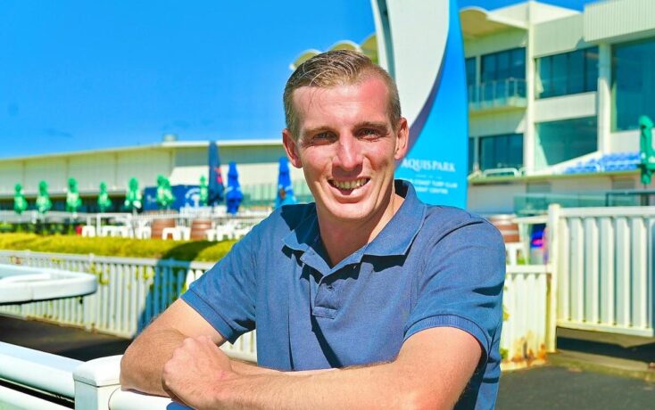 Smiling man in a blue polo leaning on a white railing at an outdoor aquatics facility on a sunny day
