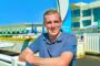 Smiling man in a blue polo leaning on a white railing at an outdoor aquatics facility on a sunny day