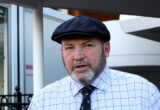 Close-up of an older man wearing a dark navy flat cap and a light, checkered shirt, outdoors beside a building.