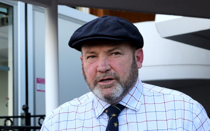 Close-up of an older man wearing a dark navy flat cap and a light, checkered shirt, outdoors beside a building.