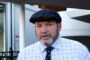 Close-up of an older man wearing a dark navy flat cap and a light, checkered shirt, outdoors beside a building.
