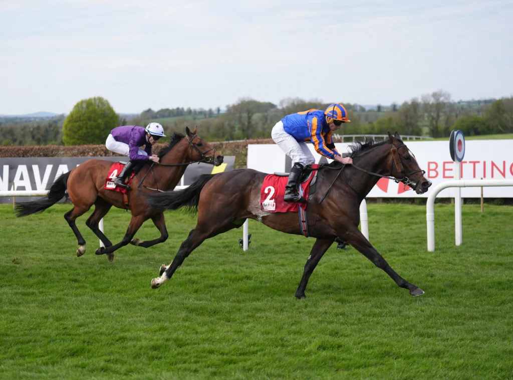 Two jockeys race side by side on horses across a grassy track, one in purple silks and the other in blue and orange, leaning forward in sprinting position.