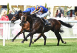 Two horses sprinting side by side on a grassy racetrack, jockeys leaning forward in bright blue and orange silks amid a cheering crowd behind a white fence.