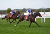 Two jockeys race side by side on horses across a grassy track, one in purple silks and the other in blue and orange, leaning forward in sprinting position.
