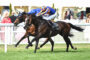 Two horses sprinting side by side on a grassy racetrack, jockeys leaning forward in bright blue and orange silks amid a cheering crowd behind a white fence.