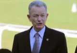 Man in a dark suit and patterned tie speaking outdoors on a sunny field.