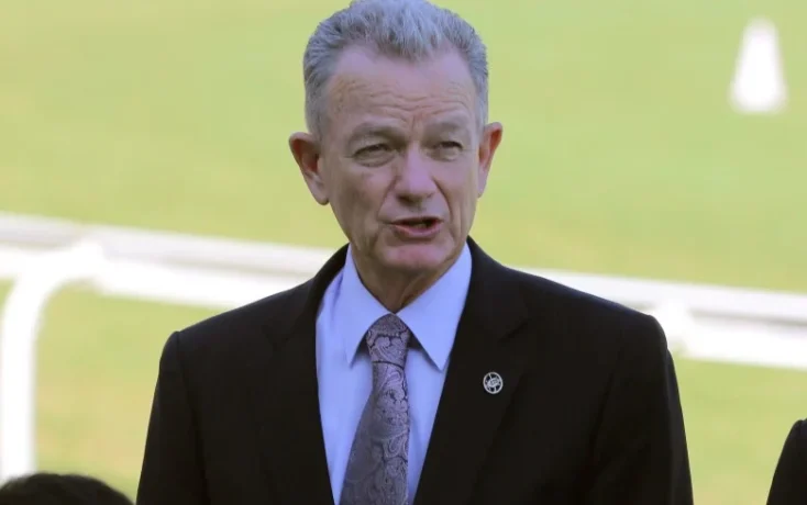 Man in a dark suit and patterned tie speaking outdoors on a sunny field.