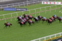 Group of thoroughbred horses and jockeys race down a green turf track, colorful silks flying as they vie for the lead with white rails and sponsor banners in the background.