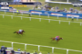Two horses race on a grassy track at Epsom Downs, with a lead horse in blue and white silks ahead of a pink-silks rival; spectators and banners in the background.