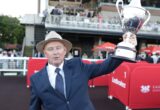 Man in a navy suit and tie wearing a straw hat raises a large silver trophy at an outdoor event, with spectators in the background.