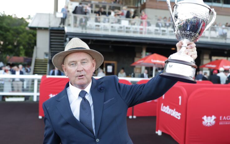 Man in a navy suit and tie wearing a straw hat raises a large silver trophy at an outdoor event, with spectators in the background.