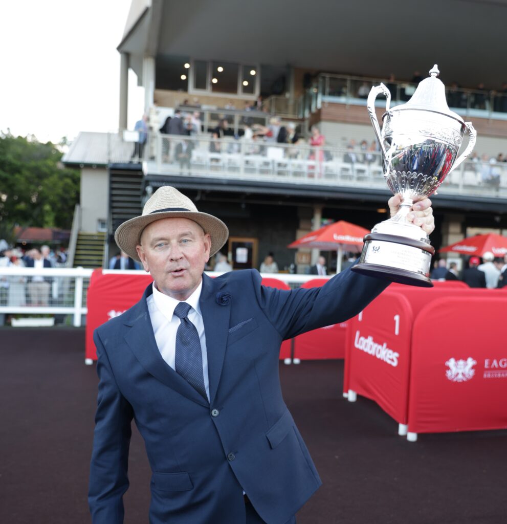 Toby Edmonds | Just Horse Racing Man in a navy suit and tie wearing a straw hat raises a large silver trophy at an outdoor event, with spectators in the background.