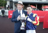 Two men posing with a large silver trophy at a horse racing event; one in a suit and hat, the other in racing silks, smiles for the camera.