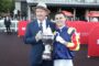 Two men posing with a large silver trophy at a horse racing event; one in a suit and hat, the other in racing silks, smiles for the camera.