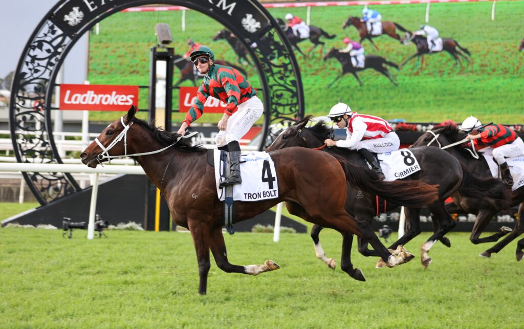 Horse race on a grassy track; jockey in red-and-green stripes rides #4 Tron Bolt ahead of rivals under a decorative start arch with Ladbrokes signs in the background.
