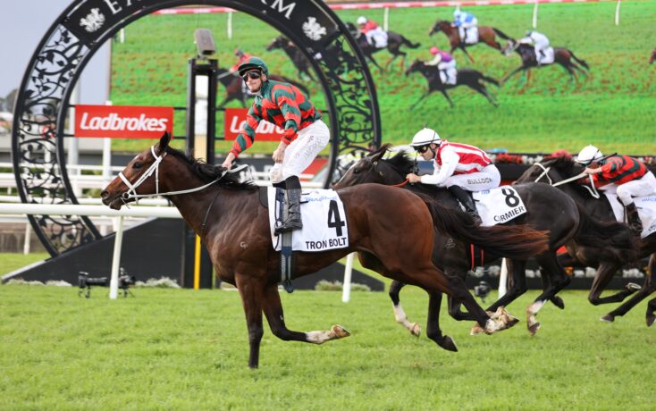Horse race on a grassy track; jockey in red-and-green stripes rides #4 Tron Bolt ahead of rivals under a decorative start arch with Ladbrokes signs in the background.