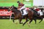 Jockey in green and red silks rides a galloping brown horse during a horse race on a green turf track, with other racers close behind and sponsor banners in the background.