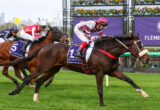Jockeys aboard galloping horses on a green turf track with a purple Flemington banner in the background.