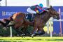 Jockey in blue helmet riding a brown racehorse sprinting on a track with Belmont Park sign in the background.