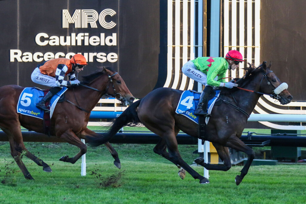 WINNASEDGEV9AWINNASEDGE889 | Just Horse Racing Two jockeys ride galloping horses in a tight race at Caulfield Racecourse, with silks in orange and green and bib numbers 5 and 4 visible on the horses' saddles.