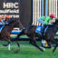 Two jockeys ride galloping horses in a tight race at Caulfield Racecourse, with silks in orange and green and bib numbers 5 and 4 visible on the horses' saddles.