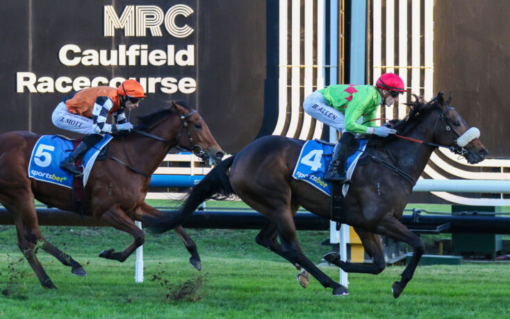 Two jockeys ride galloping horses in a tight race at Caulfield Racecourse, with silks in orange and green and bib numbers 5 and 4 visible on the horses' saddles.