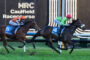 Two jockeys ride galloping horses in a tight race at Caulfield Racecourse, with silks in orange and green and bib numbers 5 and 4 visible on the horses' saddles.
