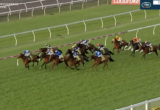 Horses and jockeys race on a green turf track with white railings and orange cones in the background.