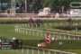 Horse race on a turf track with jockeys in colorful silks; betting odds and race info overlay in lower left corner.