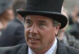 Man in a black top hat and formal suit speaking, with a blurred crowd in the background.